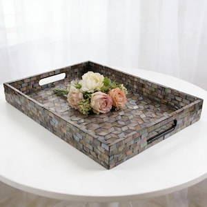May include: Rectangular decorative tray with a mosaic tile design in shades of gray, brown, and iridescent hues. The tray has built-in handles and is adorned with a bouquet of pink and cream-colored roses. The tray is placed on a white table.