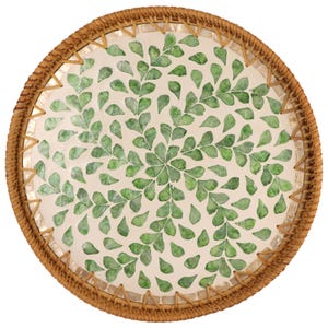 Puede incluir: Bandeja decorativa redonda con borde de ratán tejido. El centro presenta una superficie color crema adornada con un patrón de acentos verdes en forma de hojas. El borde de la bandeja está detallado con un ribete cosido de color claro.