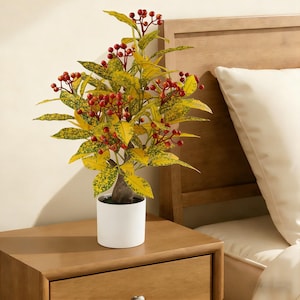 May include: A decorative artificial plant with yellow and green foliage and red berries, in a white pot. The plant is on a wooden bedside table. A wooden headboard and a white pillow are in the background.