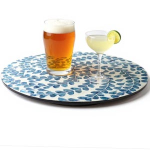 May include: A round serving tray with a blue leaf pattern holds a pint of beer and a cocktail in a coupe glass with a lime wedge. The tray has a black edge and a white background.