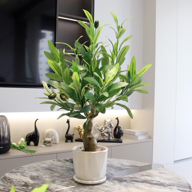 May include: A faux olive tree in a white ceramic pot. The tree has green leaves and small, dark green olives. The pot sits on a marble-patterned table. The tree is a decorative item for home decor.