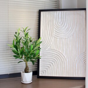May include: A decorative, framed, white textured art piece with a black frame. A small potted olive tree with green leaves and black olives sits in front of the artwork. The pot is white and geometric.
