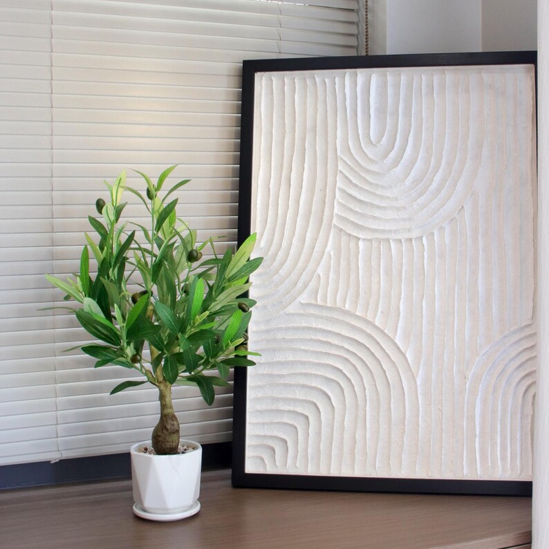 May include: A decorative, framed, white textured art piece with a black frame. A small potted olive tree with green leaves and black olives sits in front of the artwork. The pot is white and geometric.