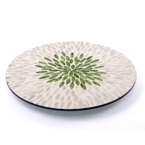 Pode incluir: Prato decorativo redondo com um design floral branco e verde. O prato apresenta um padrão central de elementos verdes em forma de folha rodeados por formas brancas em forma de pétala. O prato tem uma borda preta.