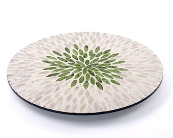 Puede incluir: Plato decorativo redondo con un diseño floral blanco y verde. El plato presenta un patrón central de elementos verdes en forma de hoja rodeados de formas blancas en forma de pétalo. El plato tiene un borde negro.
