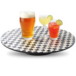 May include: A round serving tray with a black and white chequered pattern holds three drinks: a beer, a cocktail, and a red drink with a lime wedge. The tray has a black rim and is set against a white background.