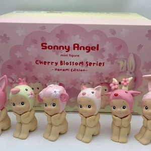 May include: A collection of six Sonny Angel mini figures from the Cherry Blossom Series, featuring adorable characters in various floral-themed hats. The figures are in a seated pose with hands clasped, presented in a pink box with matching cherry blossom graphics.