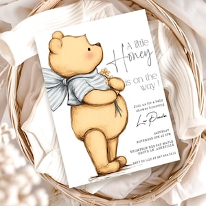 May include: A baby shower invitation featuring a watercolor illustration of a teddy bear holding flowers. The text reads "A little Honey is on the way!" in elegant script. The invitation is set in a woven basket.