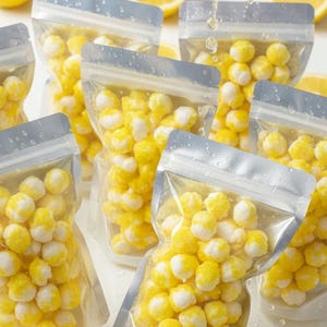 May include: A vibrant image showcasing fresh lemons and bags of yellow and white candies. The lemons are whole, sliced, and being juiced, with droplets of juice falling. The candies are in clear, sealed bags.