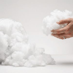 Puede incluir: Primer plano de relleno de fibra de poliéster blanca y suave, con un puñado en una mano. El relleno de fibra se asemeja a las nubes, ideal para rellenar cojines, almohadas o proyectos de manualidades. El fondo es blanco brillante.