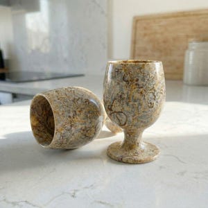 Puede incluir: Dos copas de vino de piedra con un patrón único de remolinos beige, marrón y gris. Una copa está en posición vertical, mientras que la otra está tumbada de lado. Las copas tienen un tallo y una copa redondeada, perfectas para servir vino.
