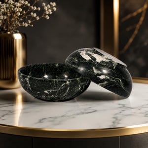 May include: Two round bowls crafted from dark green marble with prominent white veining. One bowl sits upright while the second bowl is tilted and resting against the first, showcasing the polished stone texture and natural marbled patterns.