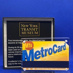 May include: A New York Transit Museum display featuring a MetroCard. The card is yellow and blue with the MTA logo and the word "MetroCard." The display includes text about the card's history.