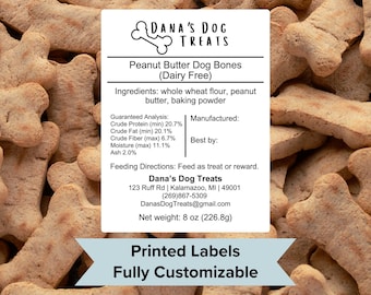 May include: A white label with the text "Dana's Dog Treats" in black. The label is for peanut butter dog bones that are dairy free. The ingredients are listed as whole wheat flour, peanut butter, and baking powder. The label also includes guaranteed analysis, feeding directions, and contact information. The label is printed on a white background and has a light blue border. The label is surrounded by a pile of dog bone shaped treats.
