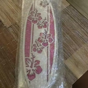May include: A surfboard-shaped decorative item with a white base and pink accents. The design features pink hibiscus flowers and vertical stripes. The item is packaged in clear plastic.