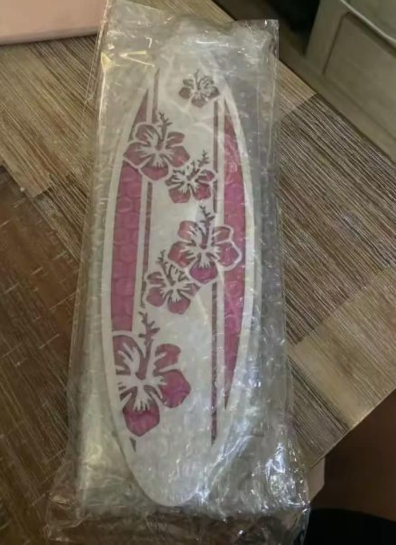 May include: A surfboard-shaped decorative item with a white base and pink accents. The design features pink hibiscus flowers and vertical stripes. The item is packaged in clear plastic.