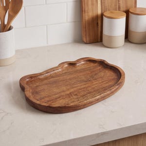 May include: A wooden serving tray with a unique, cloud-like shape. The tray is made of a warm-toned wood and has a slightly raised edge. It is sitting on a light-colored countertop. Kitchenware.