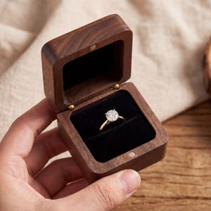 Könnte beinhalten: Geöffnete Holz-Ringbox mit einem goldenen Ring und einem klaren Edelstein auf schwarzem Samt. Die Box ist braun und hat einen Magnetverschluss. Das Schmuckkästchen ist aus Holz.