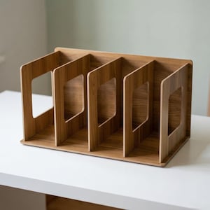 May include: A wooden desk organiser with five vertical compartments. The organiser is made of light brown wood and has a rectangular base. Each compartment has a rectangular cutout. The organiser is designed to hold files, papers, or other office supplies.