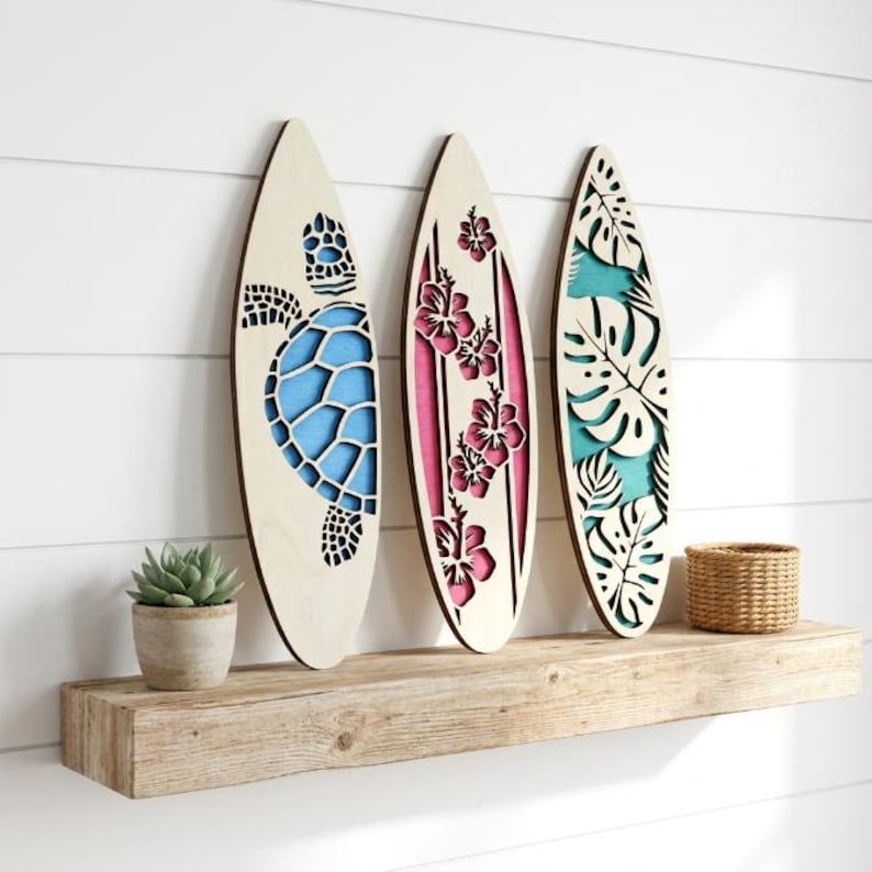 May include: Three wooden surfboards with laser-cut designs. One features a blue turtle, another pink hibiscus flowers, and the third has turquoise leaves. They are displayed on a wooden shelf with a small plant and a woven basket.