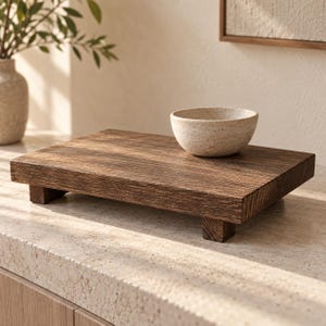 May include: A rectangular dark brown wooden riser with four short block feet, featuring a visible wood grain texture. A small, speckled off-white ceramic bowl with a smooth, rounded rim sits on the right side of the wooden platform surface.