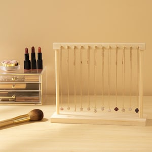 May include: A wooden jewelry display stand holding multiple gold necklaces with various pendant designs. The stand is on a light-colored surface, with makeup and a brush in the background. The necklaces have different charms, including stars and geometric shapes.