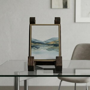 Puede incluir: Una pintura de paisaje enmarcada con un borde dorado, exhibida en un caballete de madera. La obra de arte representa una serena escena lacustre con tonos azules y dorados. El caballete es de madera oscura y tiene un dise&ntilde;o simple y moderno.