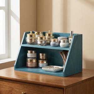 Op de afbeelding: Een turquoise houten plankeneenheid met diverse cosmetische potten en flessen. De potten hebben gouden deksels en zijn gevuld met crèmes. Een klein dienblad bevat ringen en een glas bevat make-up borstels.