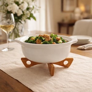 May include: A white ceramic serving bowl filled with broccoli and chicken, on a bamboo trivet. The bowl has handles and sits on a light-coloured tablecloth. A glass of wine and flowers are in the background.