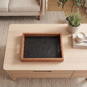 Puede incluir: Una bandeja rectangular de madera con interior negro tejido se encuentra sobre una mesa de centro de madera de color claro. La bandeja tiene un marco marrón oscuro y una superficie negra tejida texturizada. La mesa de centro está en un salón.