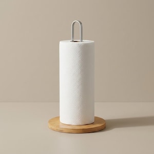 May include: A white paper towel roll with a textured surface sits on a round, light brown wooden base. A silver metal handle arches over the top of the roll. The background is a neutral beige.