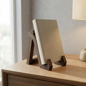 Puede incluir: Un atril de madera marr&oacute;n oscuro sostiene un libro de tapa dura con una cubierta beige. El atril tiene un dise&ntilde;o en forma de A y est&aacute; colocado sobre una superficie de madera. El libro est&aacute; en posici&oacute;n vertical, mostrando su cubierta.