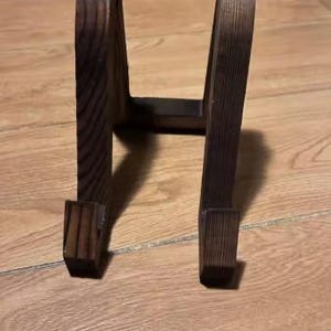 Puede incluir: Un soporte de madera marr&oacute;n oscuro con un dise&ntilde;o arqueado. El soporte tiene una base rectangular y una parte superior curvada, adecuado para exhibir objetos. La veta de la madera es visible, lo que a&ntilde;ade encanto r&uacute;stico.