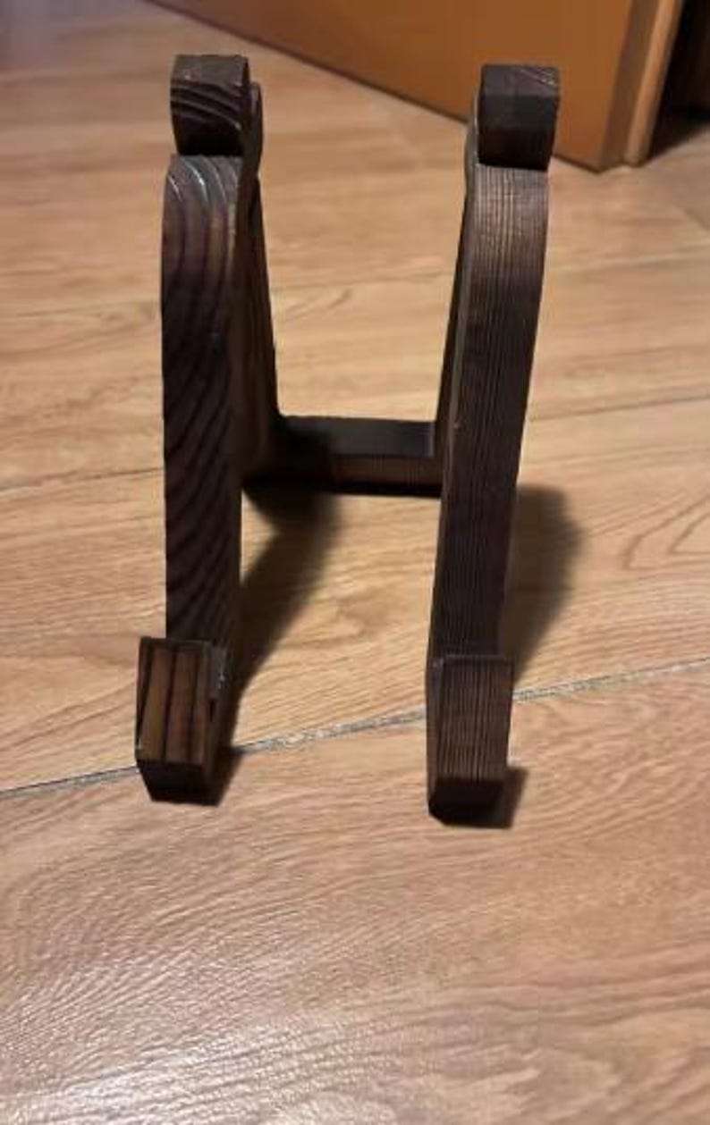 Puede incluir: Un soporte de madera marr&oacute;n oscuro con un dise&ntilde;o arqueado. El soporte tiene una base rectangular y una parte superior curvada, adecuado para exhibir objetos. La veta de la madera es visible, lo que a&ntilde;ade encanto r&uacute;stico.
