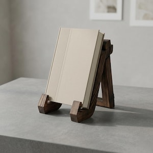 Puede incluir: Un atril de madera marr&oacute;n oscuro sostiene un libro beige. El atril est&aacute; inclinado, con una base curva y soportes. El libro est&aacute; en posici&oacute;n vertical, con una cubierta texturizada. El atril est&aacute; sobre una superficie gris.