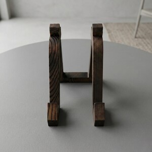 Puede incluir: Un soporte de madera marr&oacute;n oscuro con un dise&ntilde;o curvo. El soporte tiene una base rectangular y una parte superior con un dise&ntilde;o cuadrado. La veta de la madera es visible. El soporte est&aacute; sobre una superficie gris.