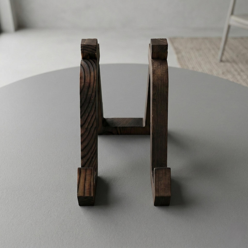 Puede incluir: Un soporte de madera marr&oacute;n oscuro con un dise&ntilde;o curvo. El soporte tiene una base rectangular y una parte superior con un dise&ntilde;o cuadrado. La veta de la madera es visible. El soporte est&aacute; sobre una superficie gris.
