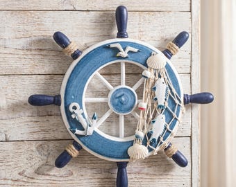 May include: A decorative nautical wall hanging depicting a ship's wheel in blue and white. It features a seagull, anchor, fish, and seashells, with rope detailing. The wheel is mounted on a light wooden panel, evoking a coastal theme.
