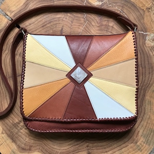May include: A brown leather shoulder bag with a geometric pattern of tan, white, and yellow leather panels. The bag has a flap closure and a decorative stone in the center.