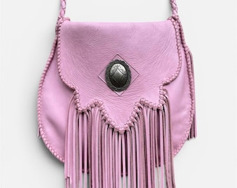 May include: A light pink leather crossbody bag featuring a braided shoulder strap, a scalloped flap edge, and long fringe detailing. A silver-toned metal concho ornament with a diamond-shaped center is centered on the front flap of the bag.