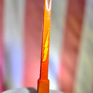 Könnte beinhalten: Ein leuchtend orangefarbenes, schmales und längliches Werkzeug mit einer gegabelten Spitze und einem rechteckigen Sockel. Der mittlere Schaft zeigt ein erhabenes, gelbes Blitzsymbol. Das Objekt weist eine texturierte, geschichtete Oberfläche auf.