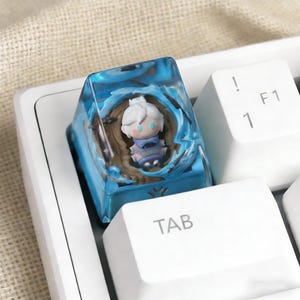 May include: A custom artisan keycap with a miniature character in blue resin. The keycap is designed for a keyboard, with the "TAB" key visible. The character has white hair and a blue outfit. The resin has a wave-like design.