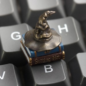 May include: A bronze-colored Sorting Hat figurine atop two blue and brown books, resting on a computer keyboard. The books have gold lettering that reads "Sorting Hat". The hat is a classic wizard's hat.