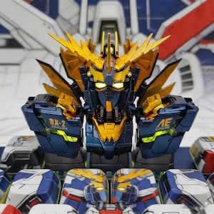 May include: Close-up of a detailed Gundam model. The robot features a gold and navy blue color scheme with intricate detailing. The text "RX-2 UNICORN" and "AE GUNDAM" are visible on the model's arms. The model has a futuristic design.