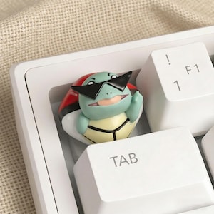 May include: A custom keycap featuring a green turtle character wearing black sunglasses, set on a white keyboard. The keycap is a novelty item, with the turtle character positioned on a red and white Pokeball.