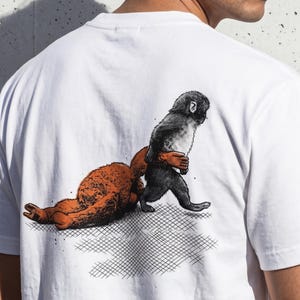 Könnte beinhalten: Weißes T-Shirt mit einer grafischen Illustration von zwei Cartoon-Kreaturen. Eine ist orange und liegt, die andere, in Schwarz und Grau, scheint sie zu ziehen. Das Design ist auf der Rückseite des T-Shirts aufgedruckt.