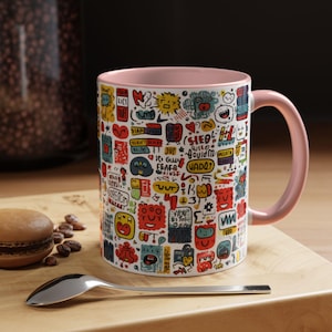 Puede incluir: Una taza de cerámica blanca con interior y asa rosas. La taza está cubierta de garabatos y texto de dibujos animados de colores. Una cuchara y un macaron están sobre una superficie de madera.