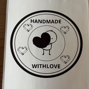 Può includere: Un adesivo circolare con bordo nero e sfondo bianco. I testi "HANDMADE" e "WITH LOVE" sono in nero. Un omino stilizzato tiene in mano un cuore nero, circondato da tre cuori.