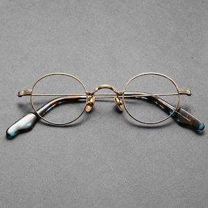 May include: Round spectacles featuring a thin, metallic gold-toned frame with intricate etched detailing on the bridge and hinges. The temples transition into curved, translucent tortoiseshell-patterned acetate tips with a dark blue hue.
