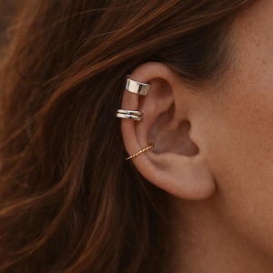 Pode incluir: Close-up de uma orelha adornada com três peças de joalheria. Uma pulseira de prata fica no topo, um brinco de prata de banda dupla no meio e um aro dourado torcido na parte inferior. A orelha está contra um fundo desfocado.